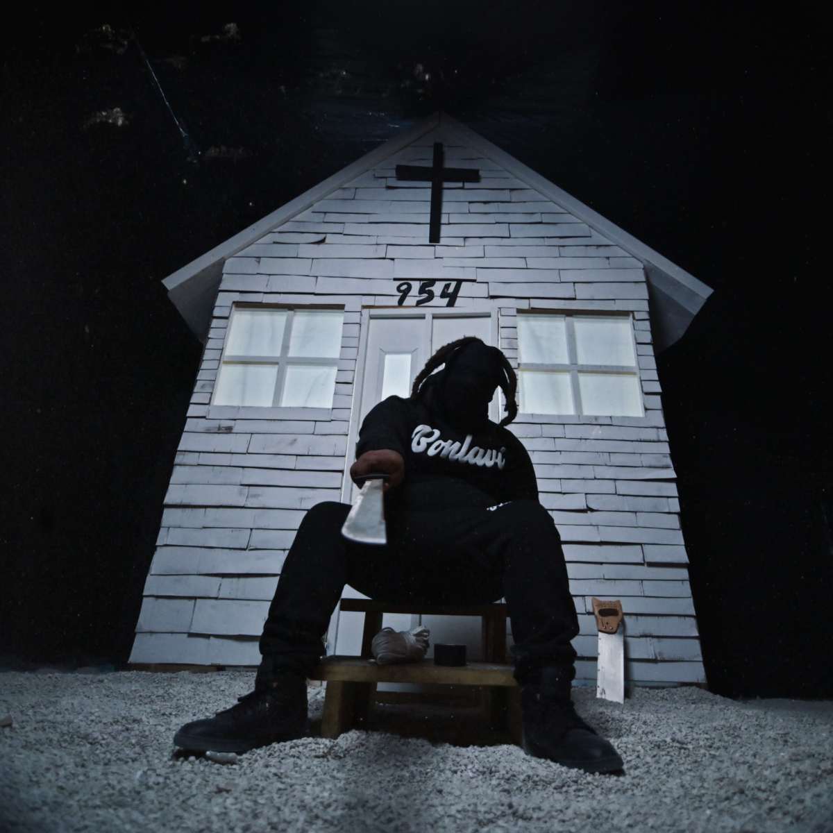 A figure in dark clothing sits on a wooden stool in front of a small, white house with a cross and the number 254 on it.