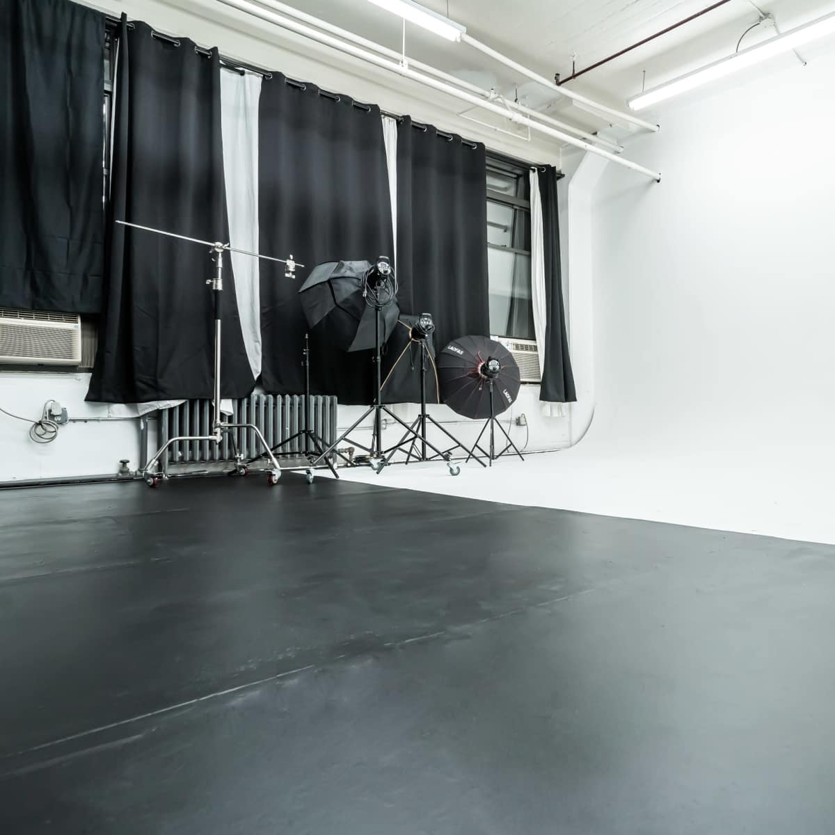 An empty photography studio with black flooring, backdrop curtains, and lighting equipment positioned in the corner.