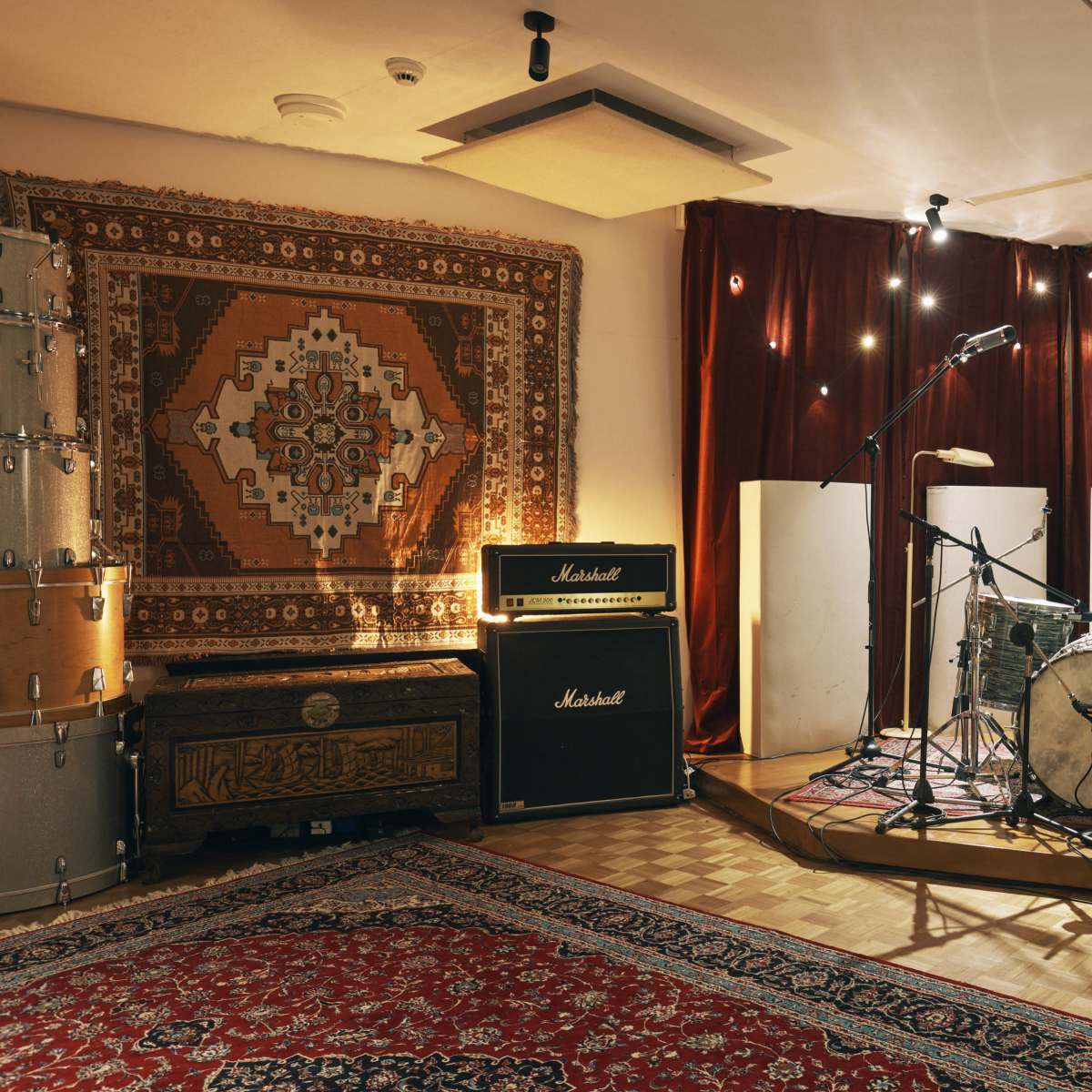 A recording studio is configured with a drum set, amplifiers, and acoustic panels, set against a backdrop of decorative rugs and soft lighting.