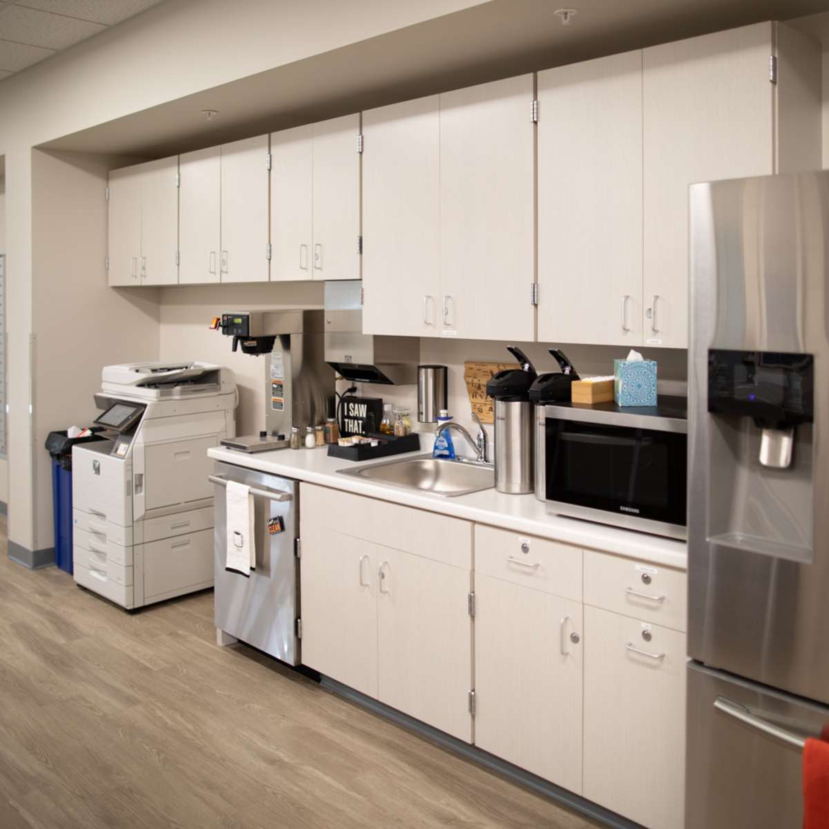 A modern office kitchen area with cabinets, a sink, a coffee machine, a microwave, and a stainless steel refrigerator.