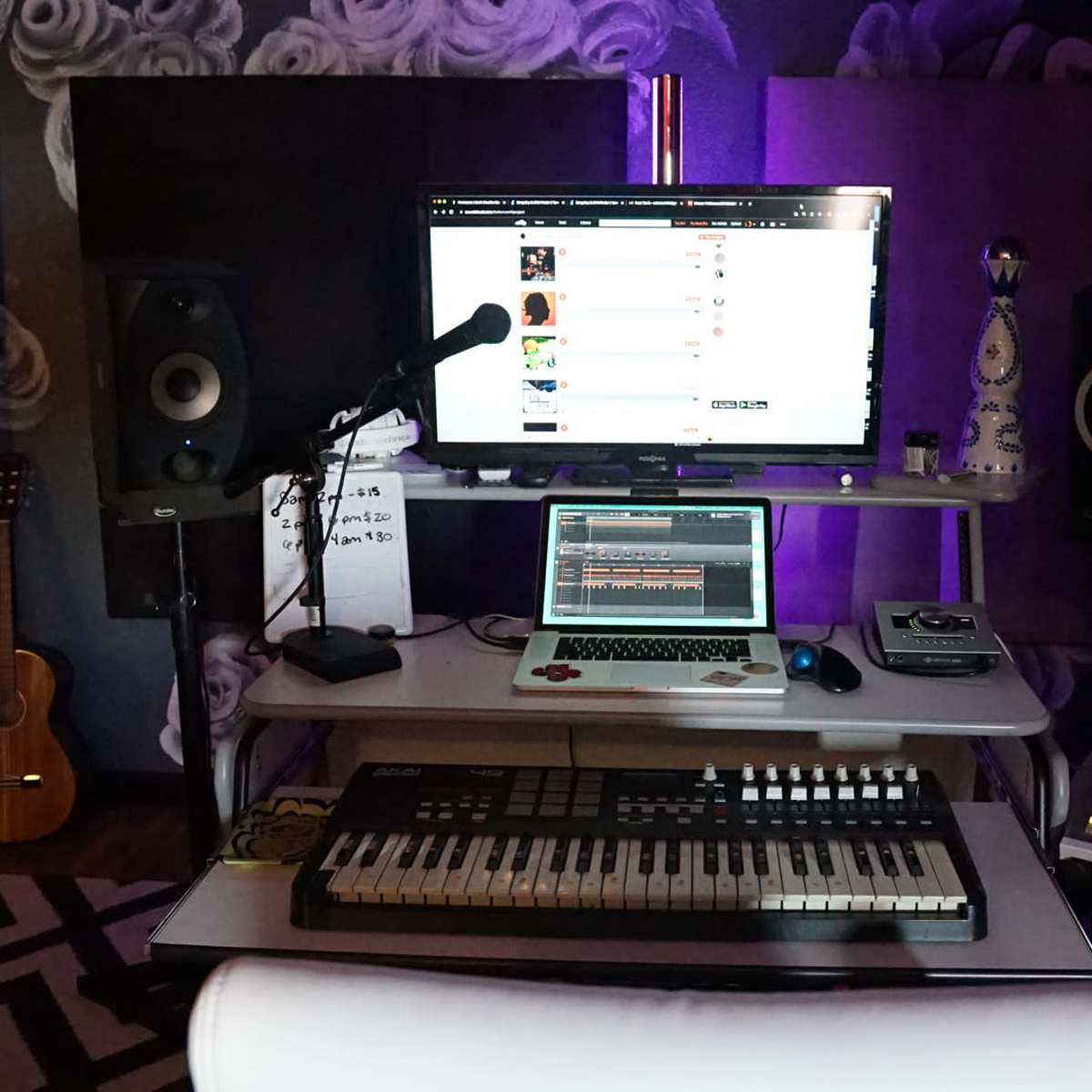 A music production setup features a computer monitor displaying a playlist, a laptop, a MIDI keyboard, and studio speakers in a dimly lit room.
