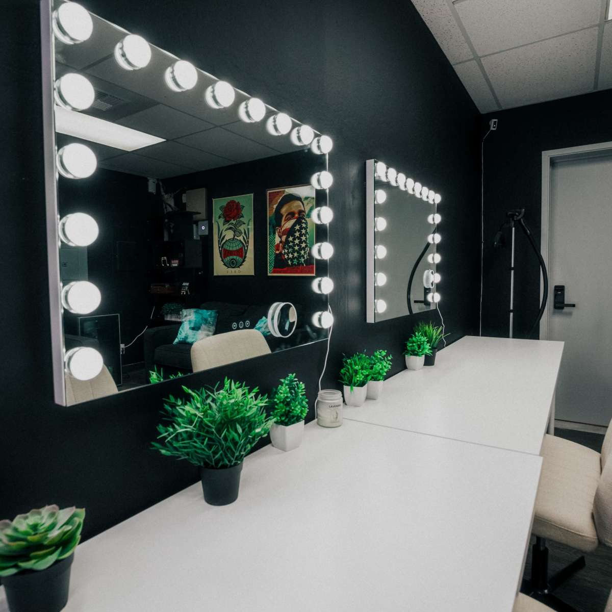 A room with two illuminated vanity mirrors above white tables, complemented by small potted plants and a door leading to another space.
