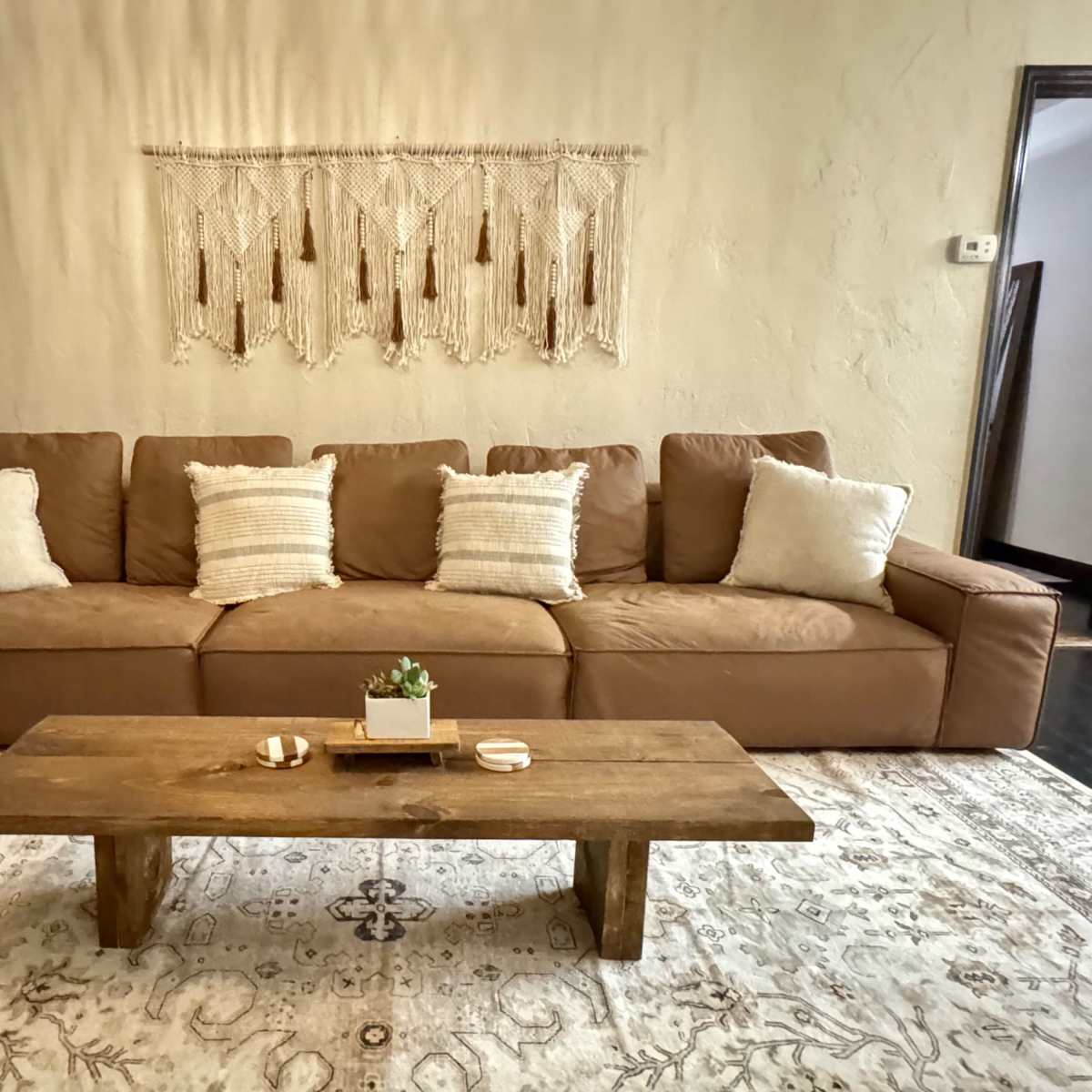 A cozy living room with a brown sectional sofa, a rustic wooden coffee table, and a decorative wall hanging above the sofa.