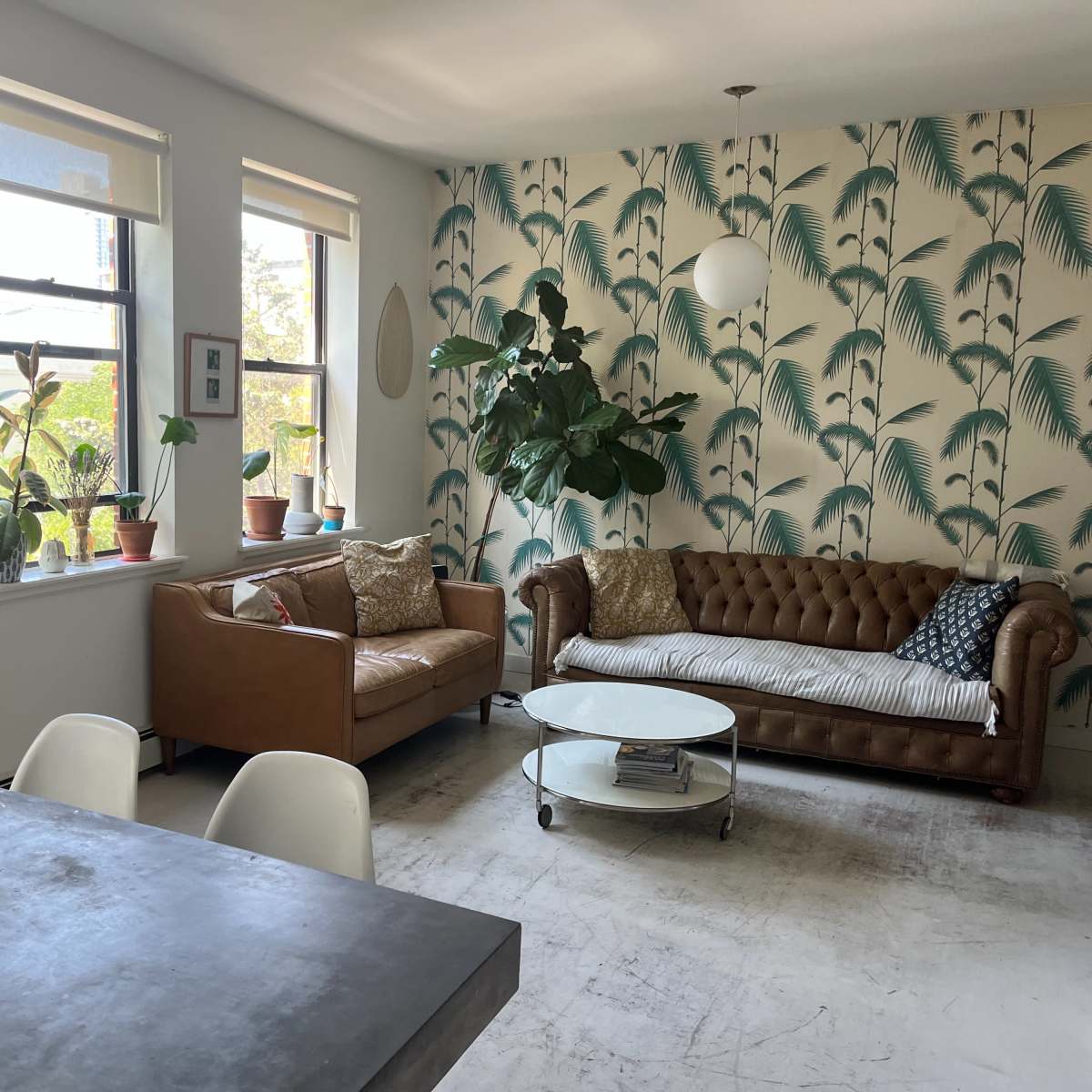A living room featuring a brown leather couch, a white coffee table, large windows with plants, and a wall decorated with tropical-themed wallpaper.
