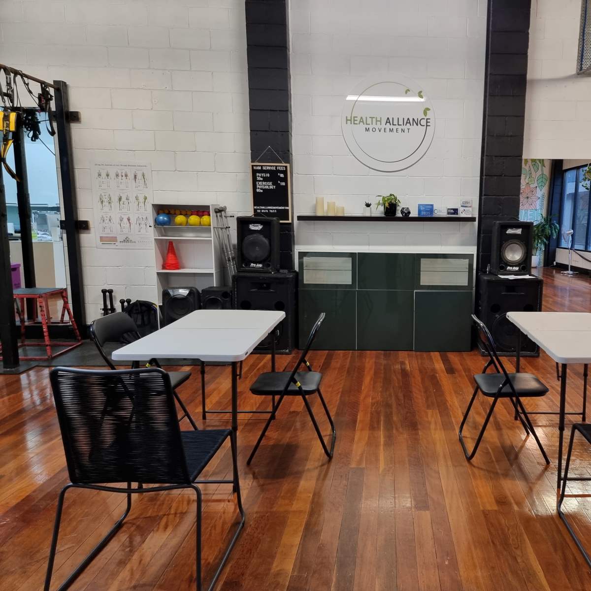 A fitness studio with a reception area featuring a round logo for "Health Alliance," surrounded by several tables and chairs in a well-lit space with wooden flooring.