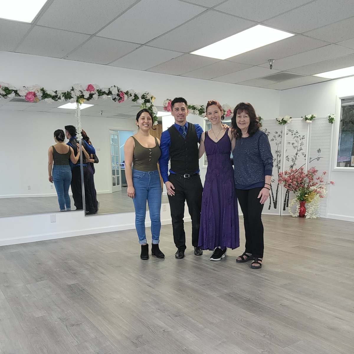 Three individuals stand in a bright dance studio with mirrored walls and floral decorations.