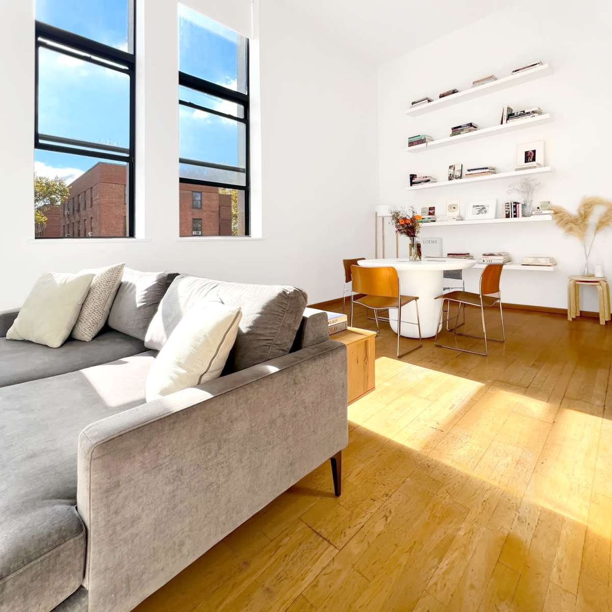 A bright, modern living space featuring a gray sofa, a small dining table with chairs, and shelves displaying books and decor against a white wall.