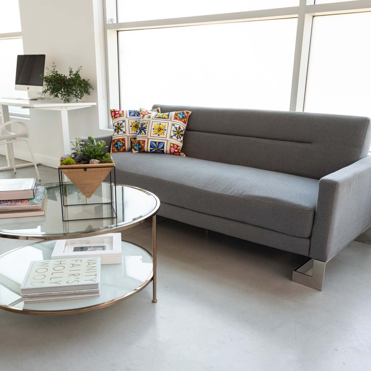 A modern living space featuring a gray sofa, a round glass coffee table with books and a plant, and a desk with a computer in the background.