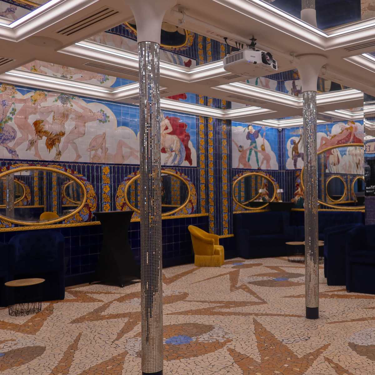 An ornate room featuring mosaic floors, blue tiled walls adorned with decorative murals, and plush seating arrangements highlighted by mirrors and unique lighting.