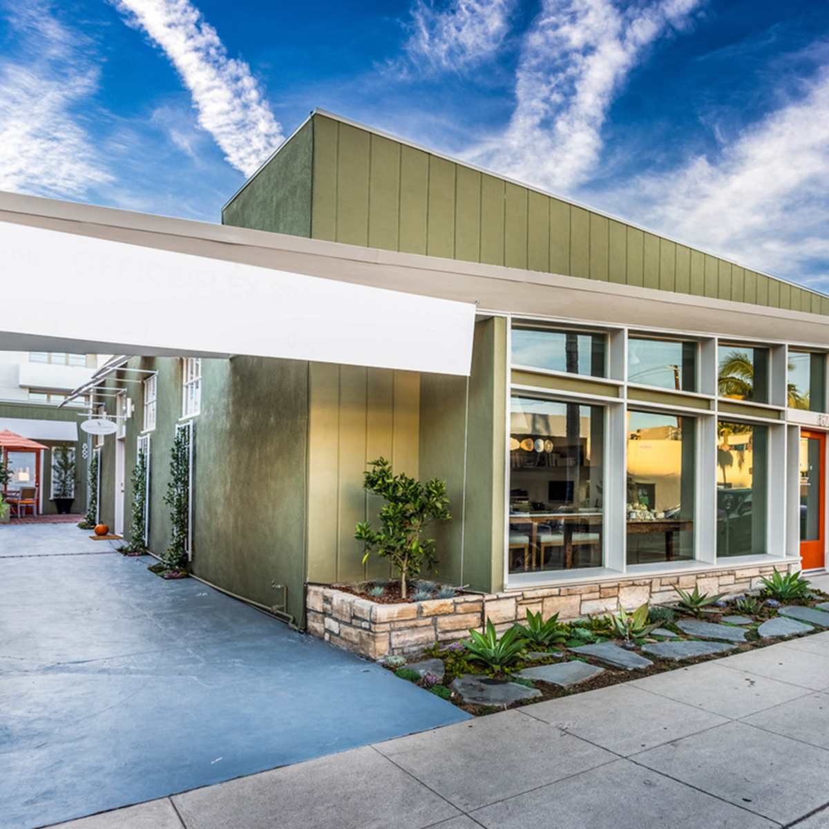 A modern single-story building with large windows, a concrete path, and landscaped areas, under a blue sky with streaky clouds.