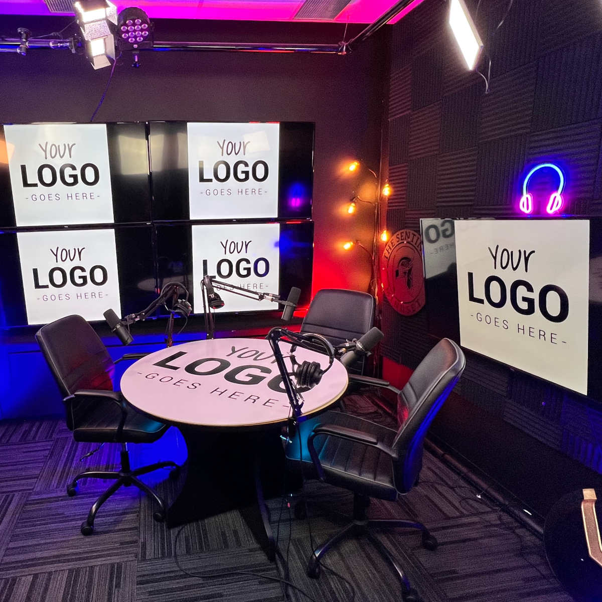 A podcast studio with a circular table, two black chairs, and multiple screens displaying the text "YOUR LOGO GOES HERE."