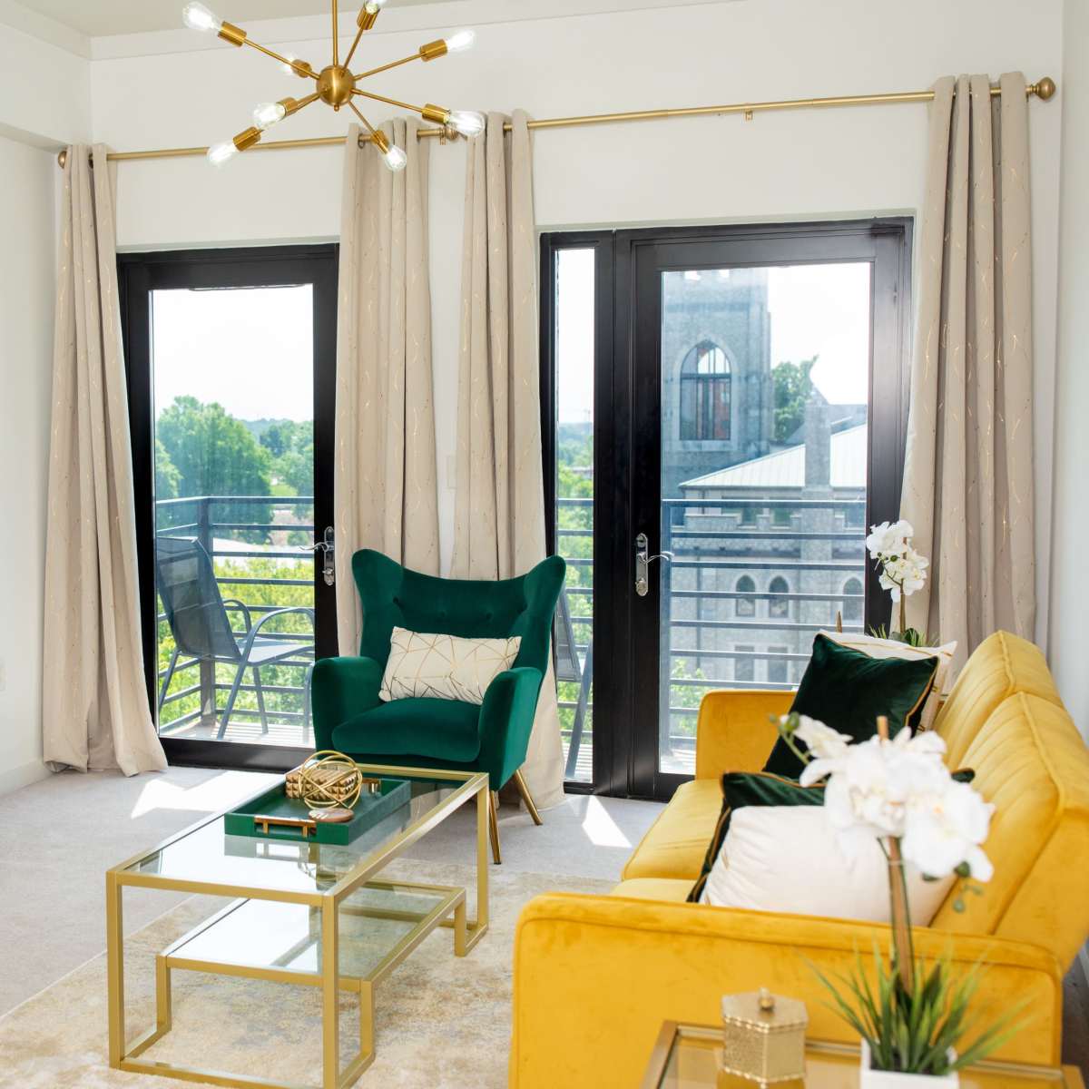 A modern living room featuring a green chair, a yellow sofa, a glass coffee table, and a television on a wooden stand, with large windows overlooking a balcony.
