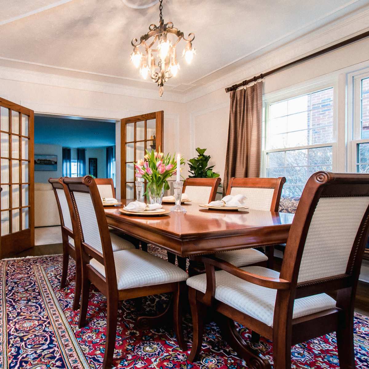 A wooden dining table is set for a meal, surrounded by upholstered chairs, in a room illuminated by a chandelier, with French doors opening to another space.