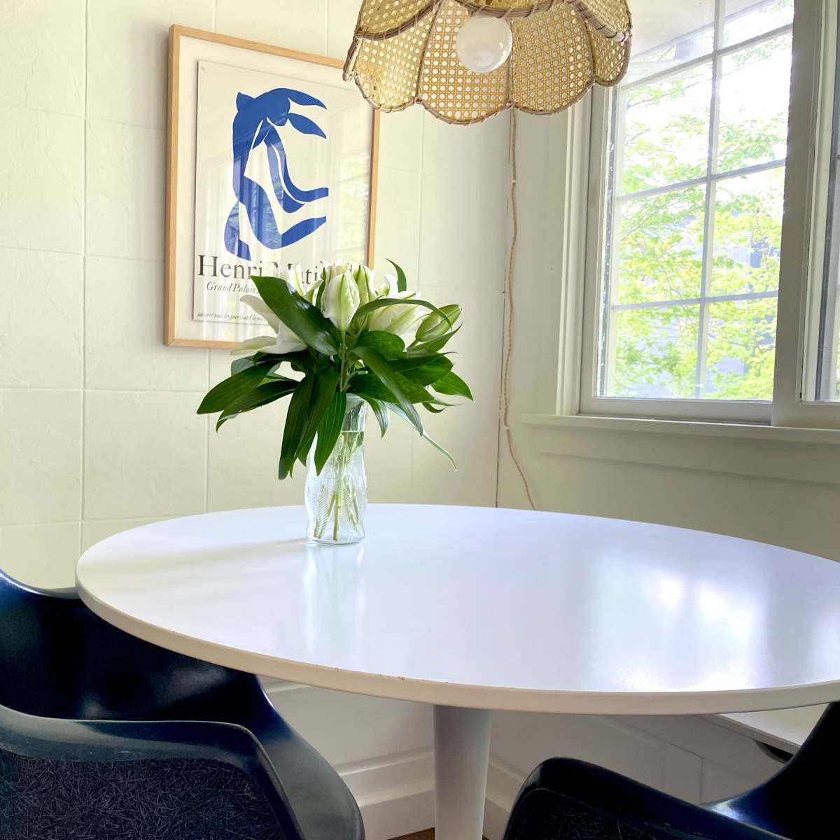 A round white table with a vase of flowers is positioned near a window, surrounded by blue and green chairs and a framed artwork on the wall.