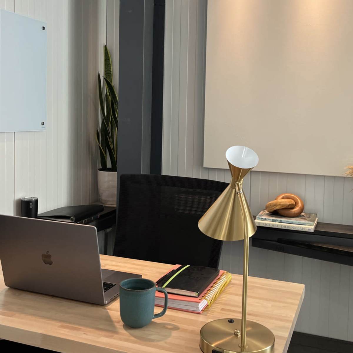 A modern office space features a wooden desk with a laptop, a coffee mug, books, a gold desk lamp, and decorative items on the wall shelf.