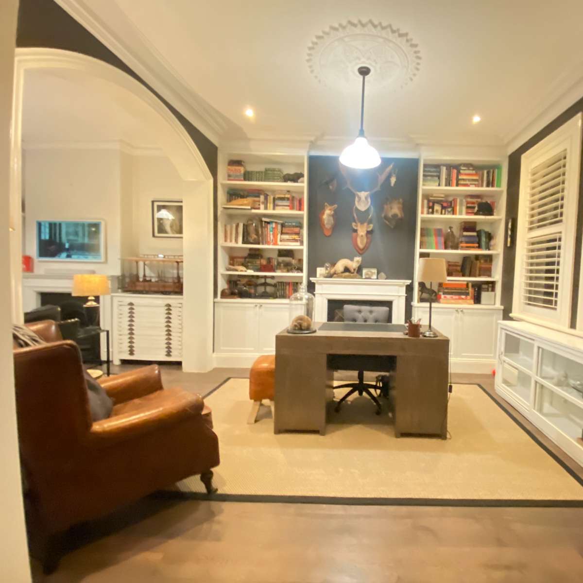 A stylish home office featuring a wooden desk, leather armchair, built-in bookshelves filled with books and decorative items, and a decorative wall with animal heads mounted.