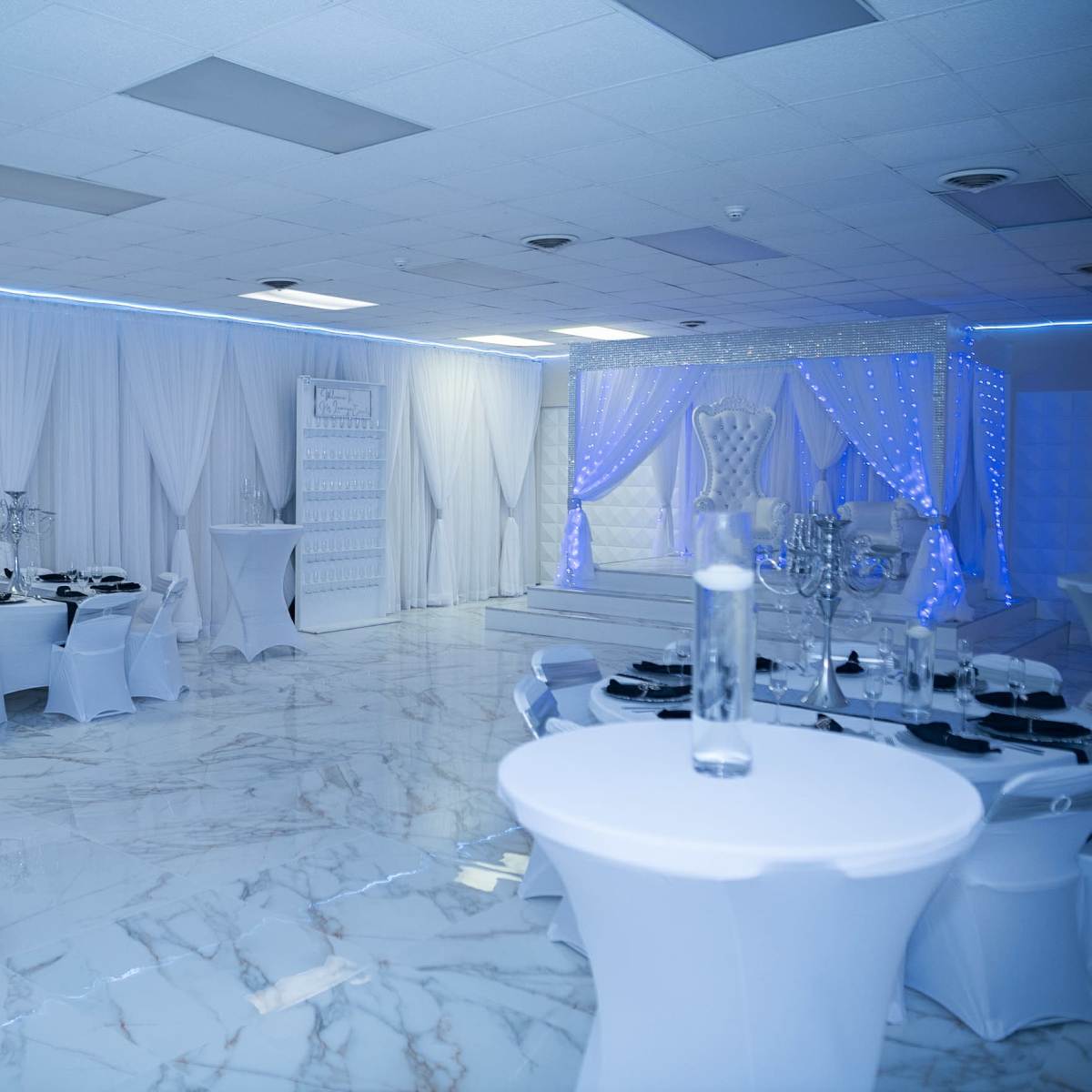 A spacious banquet hall with white drapes, marble flooring, and elegantly arranged tables set for an event.