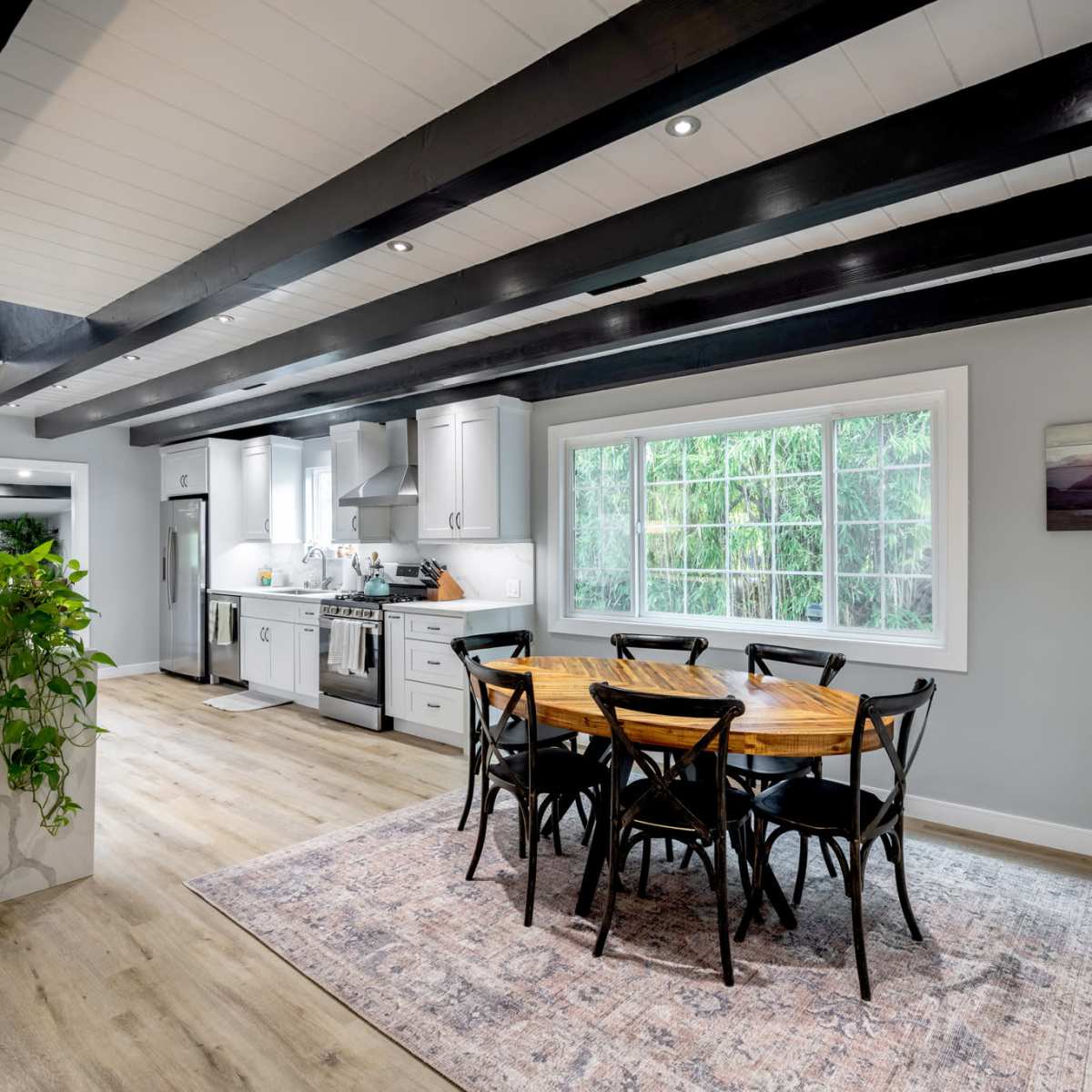 A modern kitchen and dining area featuring a wooden dining table surrounded by black chairs, with a large window allowing natural light and views of greenery outside.