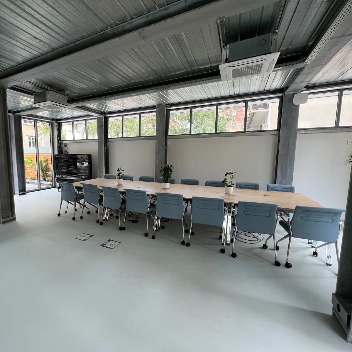 A modern meeting room features a long wooden table surrounded by blue chairs, with large windows and an elevator in the background.