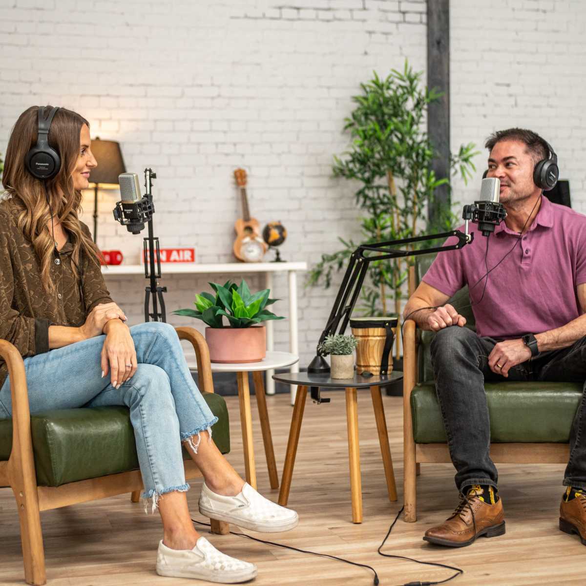 Two people are seated in green chairs in a well-lit indoor studio setup, equipped with microphones and surrounded by plants and decorative items.