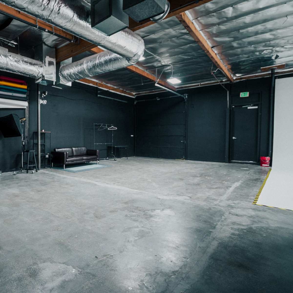 A spacious, empty studio with black walls, exposed metal ducts, and a couch along with various equipment on one side.