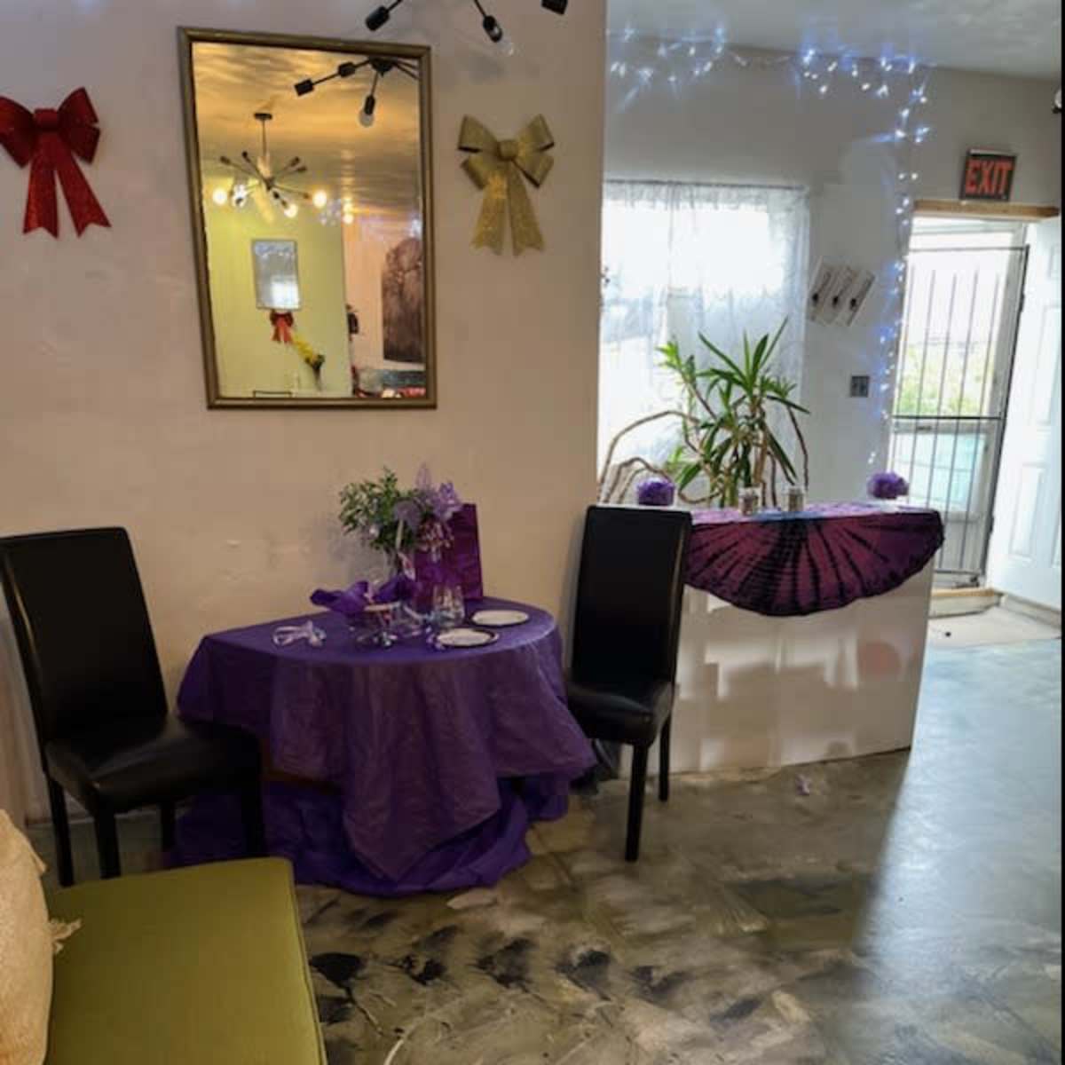 A small, decorated room with a purple tablecloth on a round table, two black chairs, and a plant near the entrance.