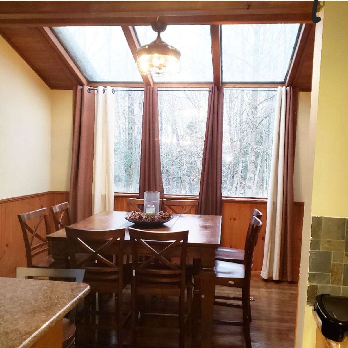 A wooden dining table surrounded by chairs is positioned in a sunlit room with large windows and curtains.