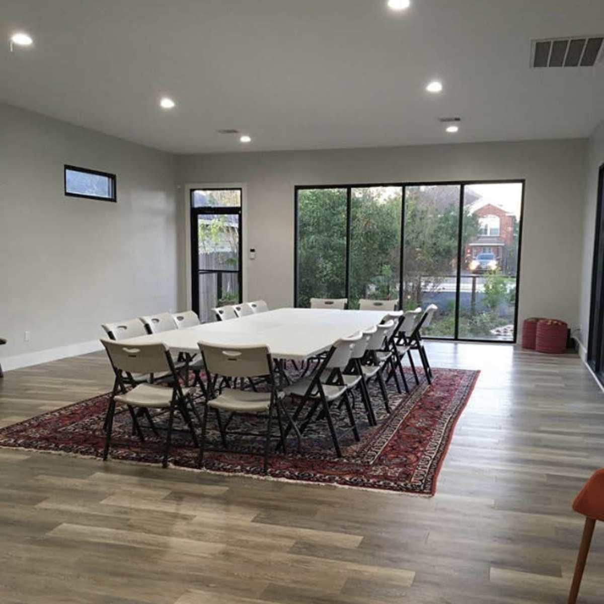 A spacious room features a long table surrounded by folding chairs, with large windows offering a view of greenery outside.