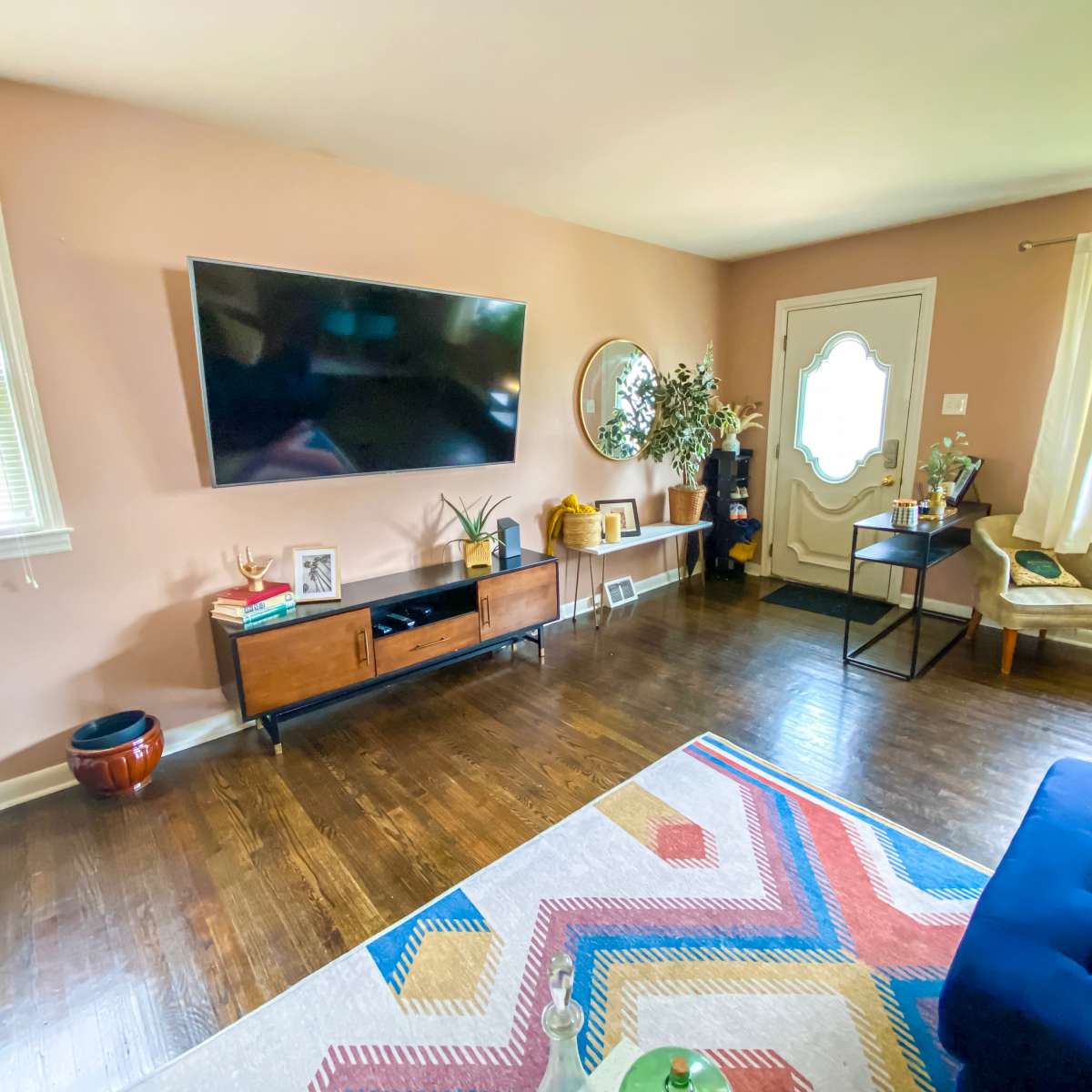 A living room features a mounted television, a wooden media console, a round mirror, a door with a window, and a colorful area rug on a hardwood floor.