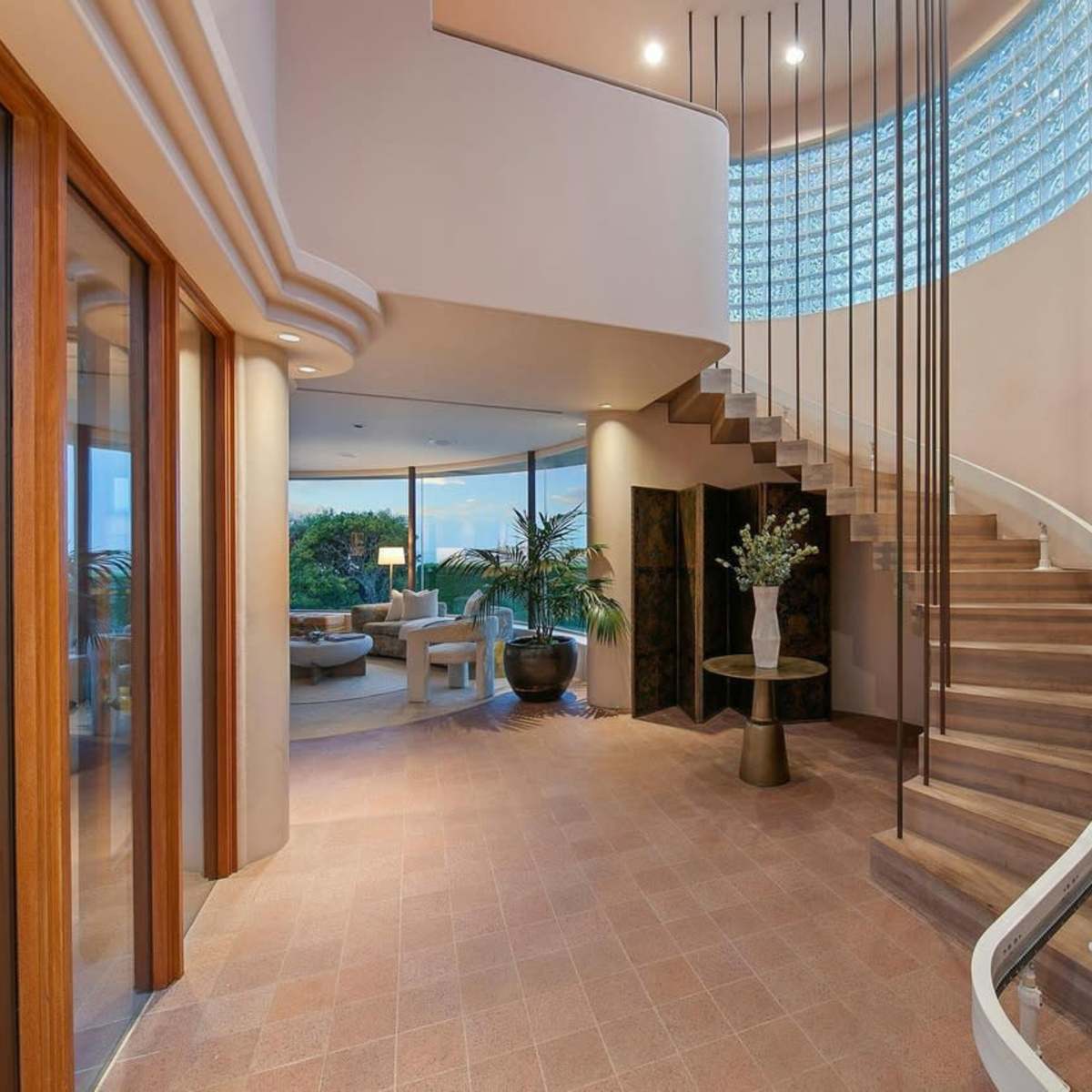 A modern entryway with a curved staircase, large glass windows, and plants arranged around the space.