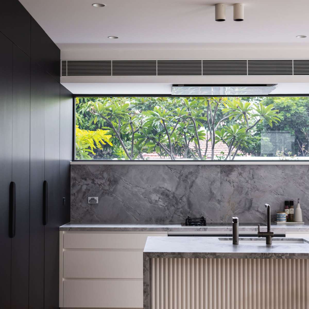 A modern kitchen with a central island, featuring a marble countertop and a large window overlooking greenery.