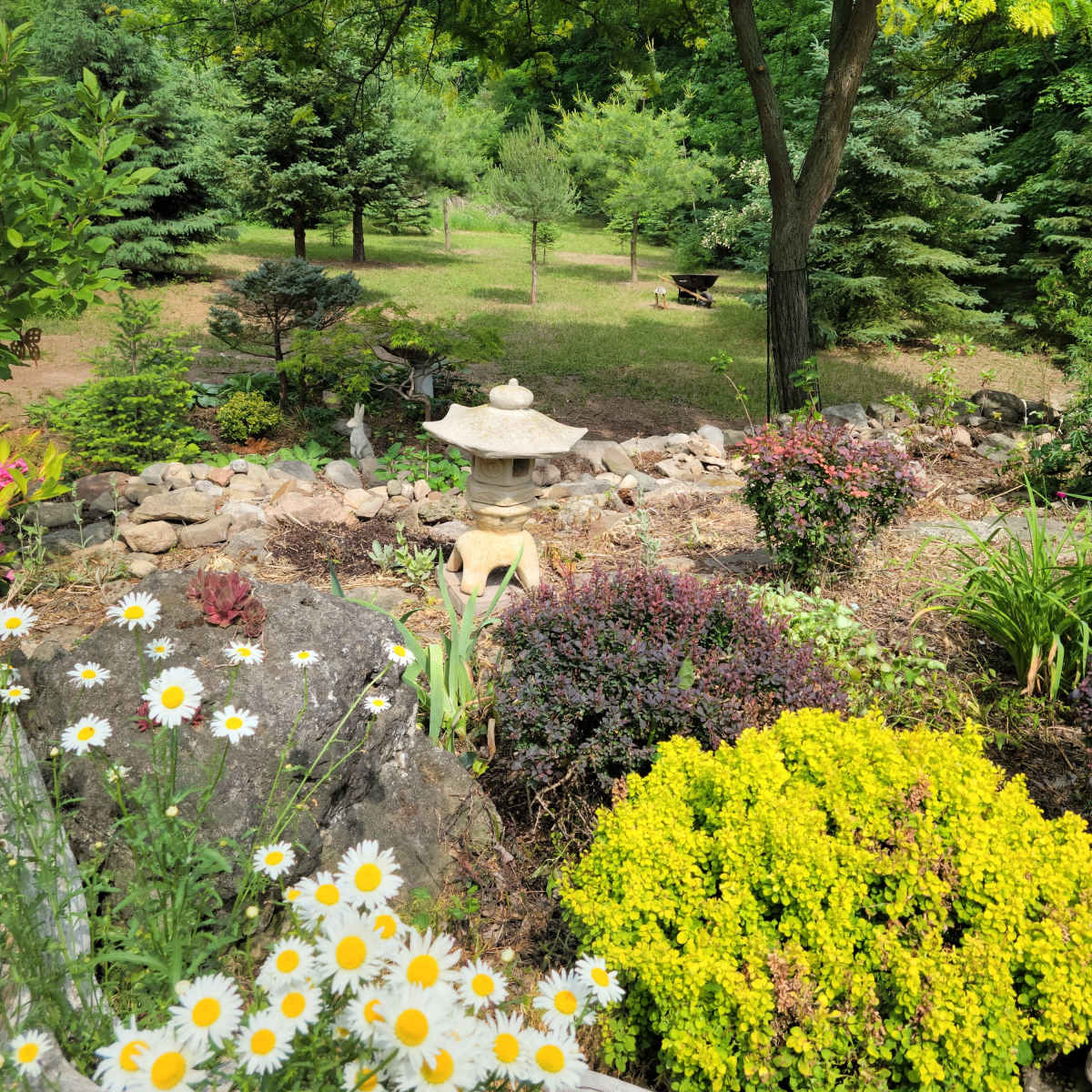 A stone lantern is surrounded by colorful flowers and shrubs in a landscaped garden featuring various trees and a grassy area in the background.