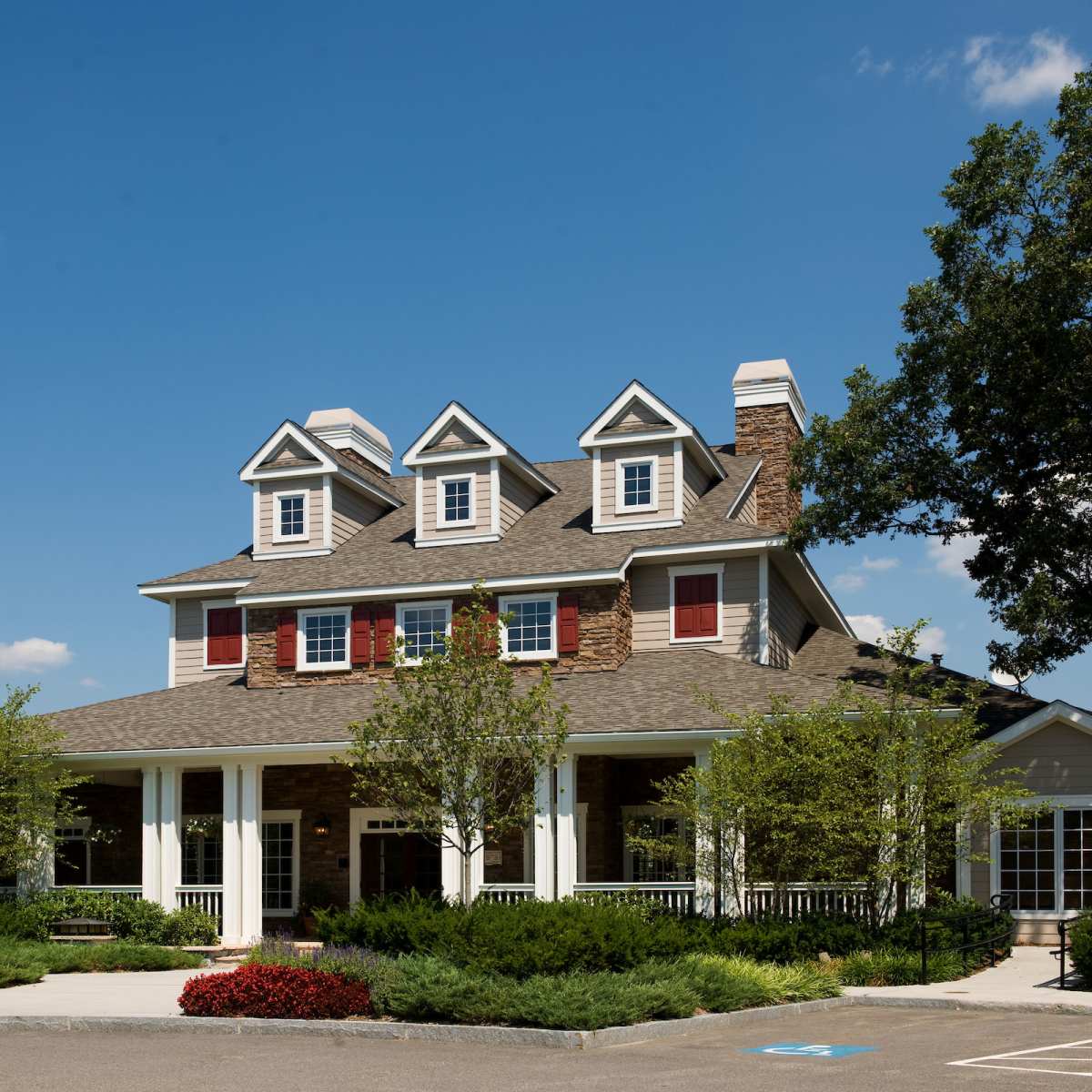 A large, multi-storied house with a stone exterior, gabled roof, and red window shutters is surrounded by neatly trimmed shrubs and trees.