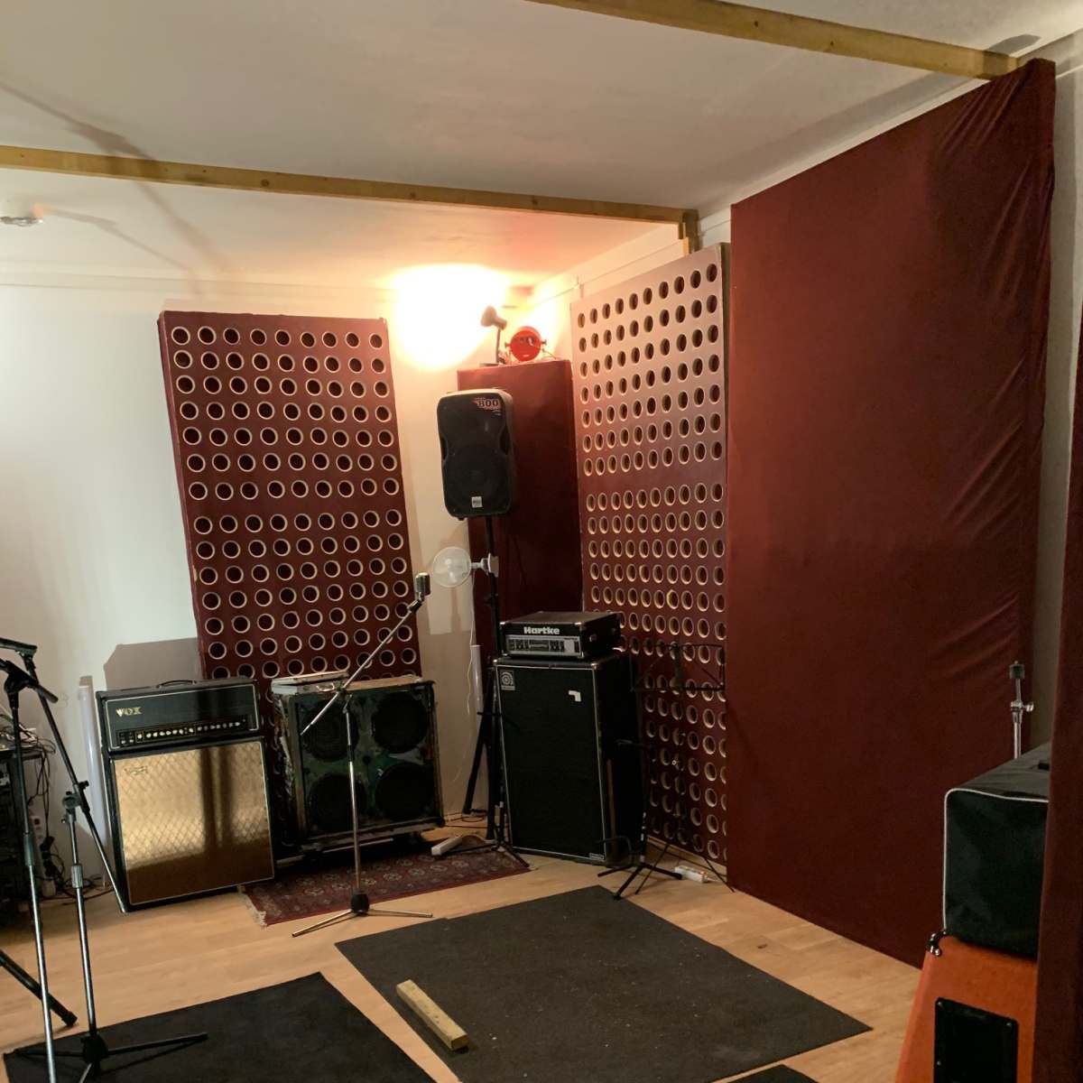 A music rehearsal space with sound-dampening panels, amplifiers, and microphones arranged around a wooden floor.