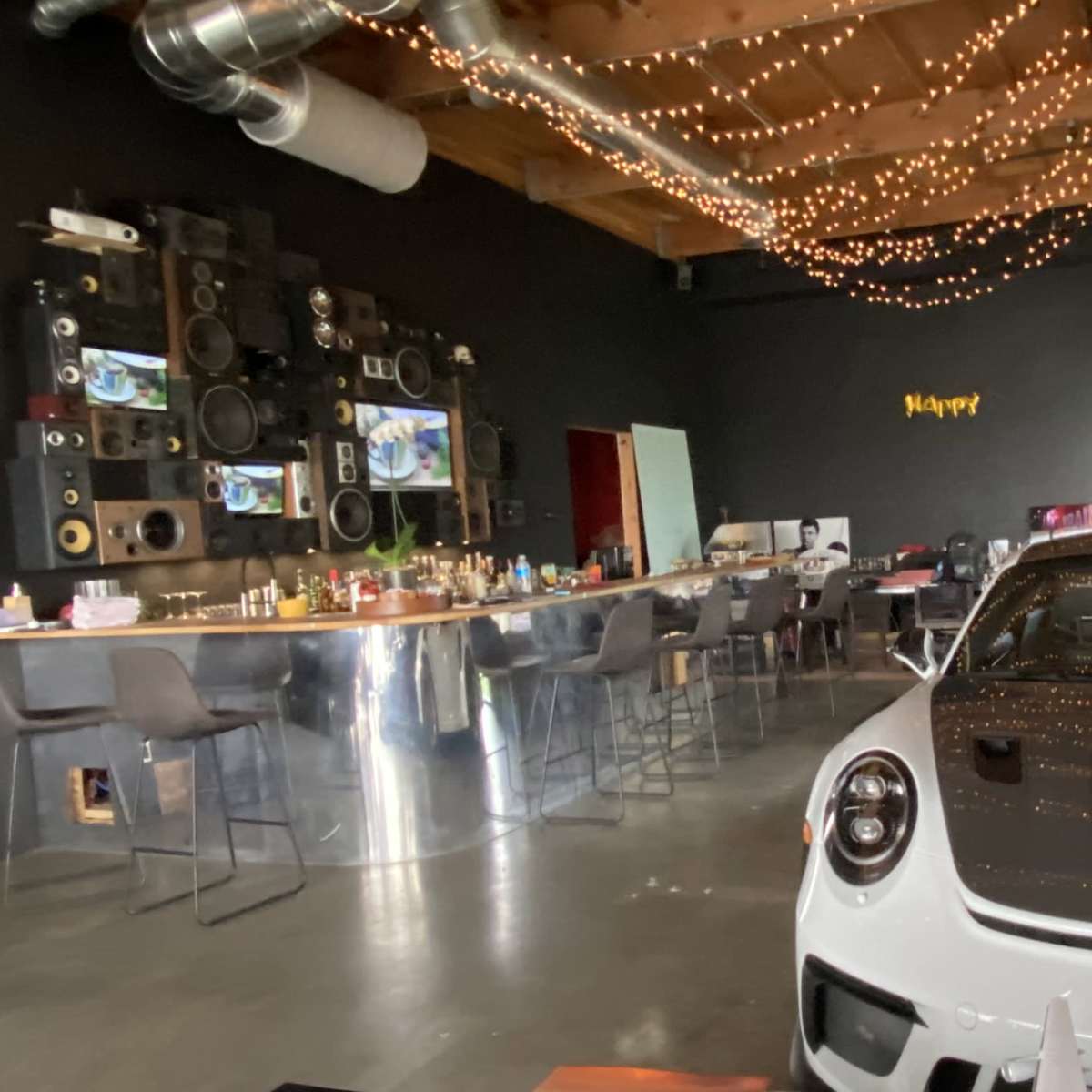 A white Porsche sports car is parked in a modern, stylish room featuring a long bar, high stools, and a wall adorned with large speakers and fairy lights.