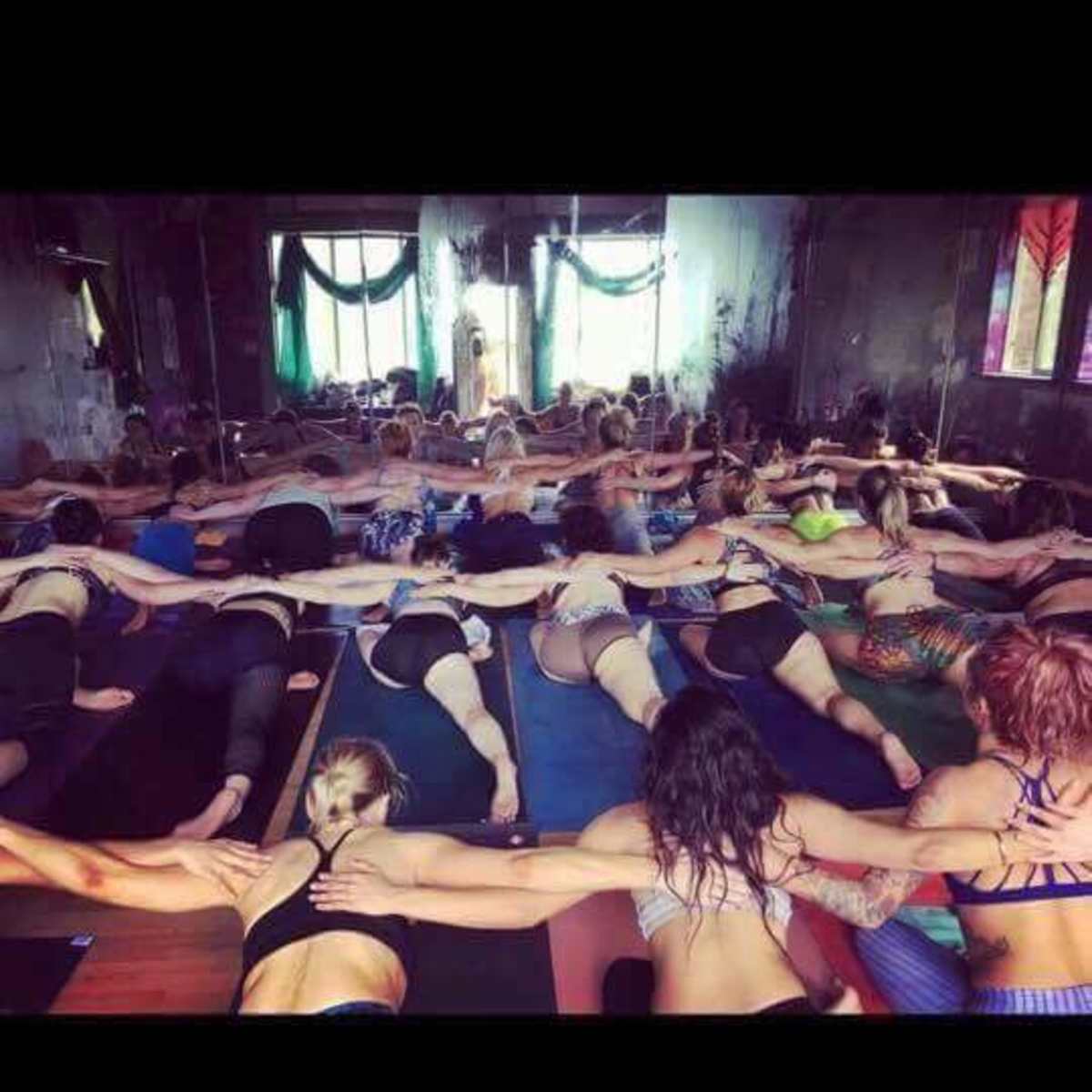 A large group of people practice yoga in a dimly lit studio, connected by holding hands while lying on mats.