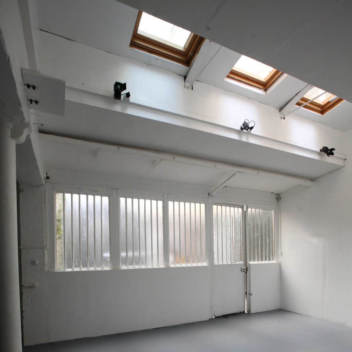 A spacious, empty room with white walls, high ceilings, and windows with vertical slats allowing light to enter.
