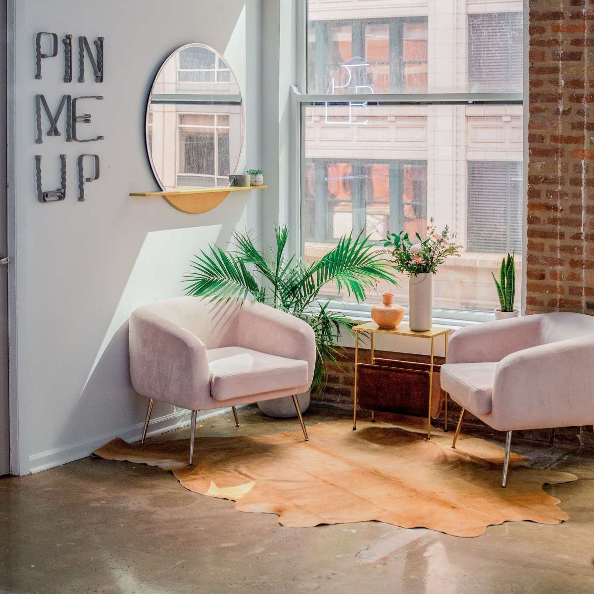 A modern seating area features two light pink armchairs, a wooden coffee table, and a round mirror, with plants and a decorative vase beside a large window.