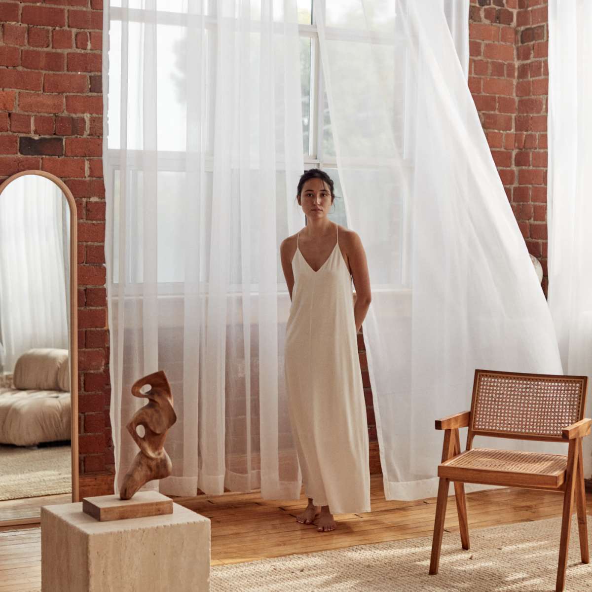 A woman stands by large, sheer curtains in a room with exposed brick walls and minimal furnishings.