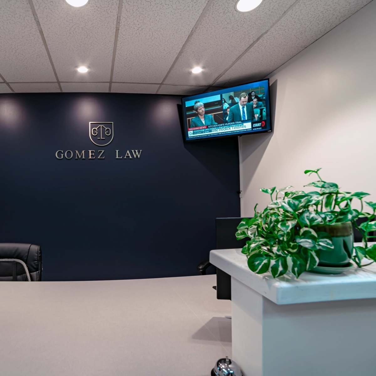 A law office reception area with a dark blue wall featuring the logo "GOMEZ LAW," a television displaying news, and potted plants on the reception desk.