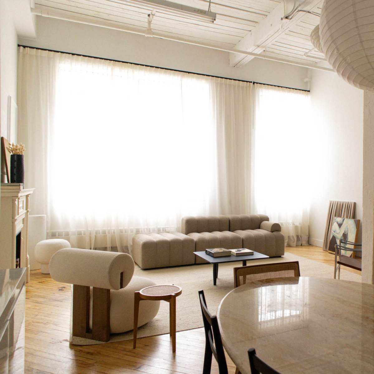 A spacious living area features large windows with sheer curtains, a sectional sofa, a mirror, a dining table, and minimalist furniture.