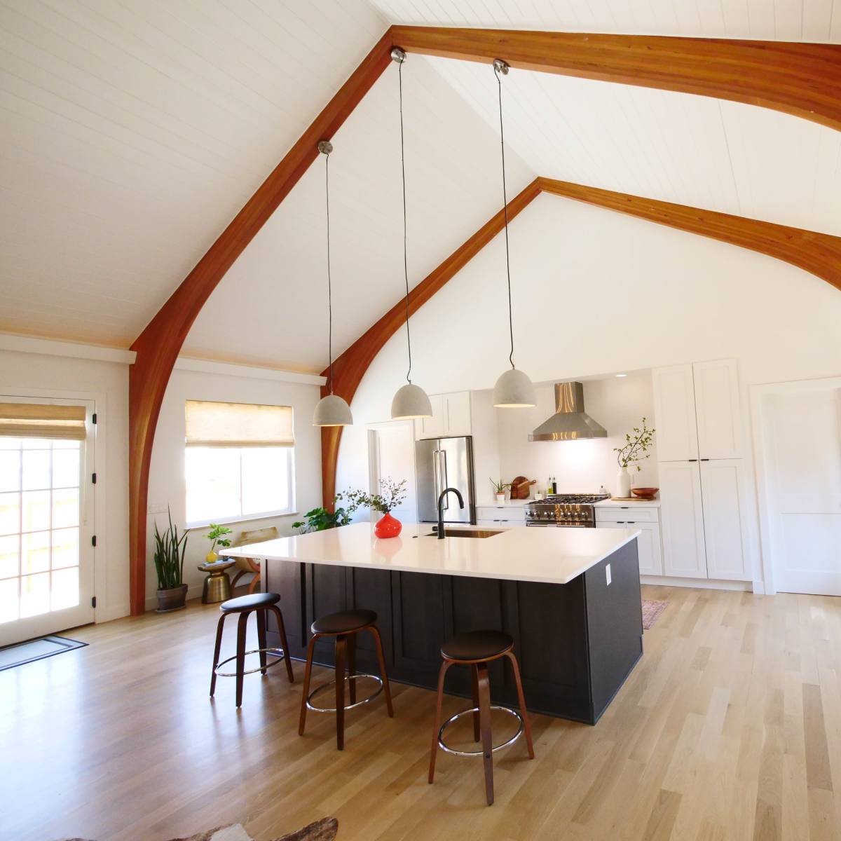 A modern kitchen with an island, wooden beams, and large windows, featuring a variety of plants and pendant lighting.
