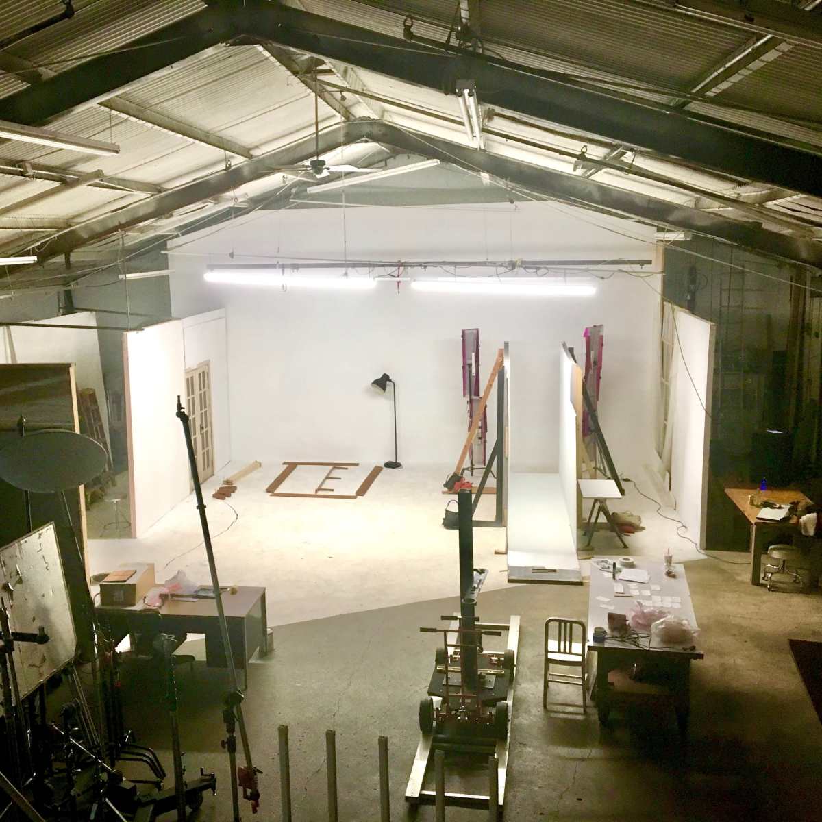 An industrial-style studio space with a white backdrop, various equipment, and scattered furniture, illuminated by overhead lights.