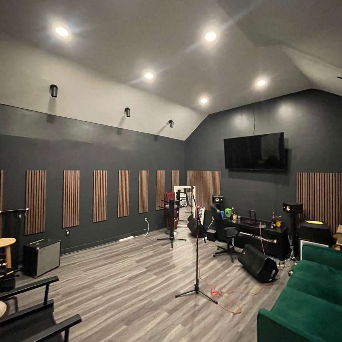 A music rehearsal studio featuring a microphone stand, seating, and sound-absorbing wall panels, with a television mounted on the wall.