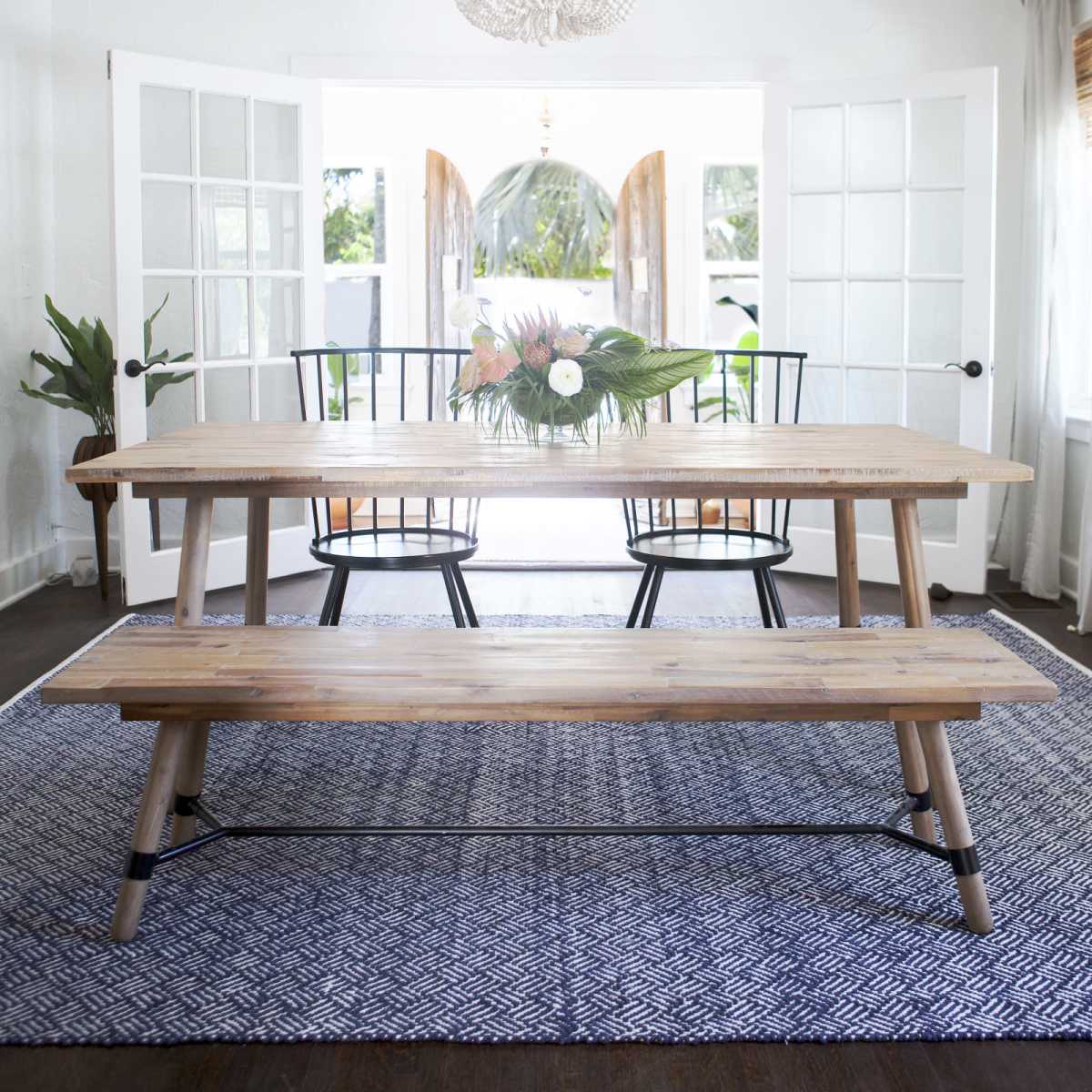 A wooden dining table with a floral centerpiece and black chairs is set in a bright room with open French doors leading to an outdoor view.