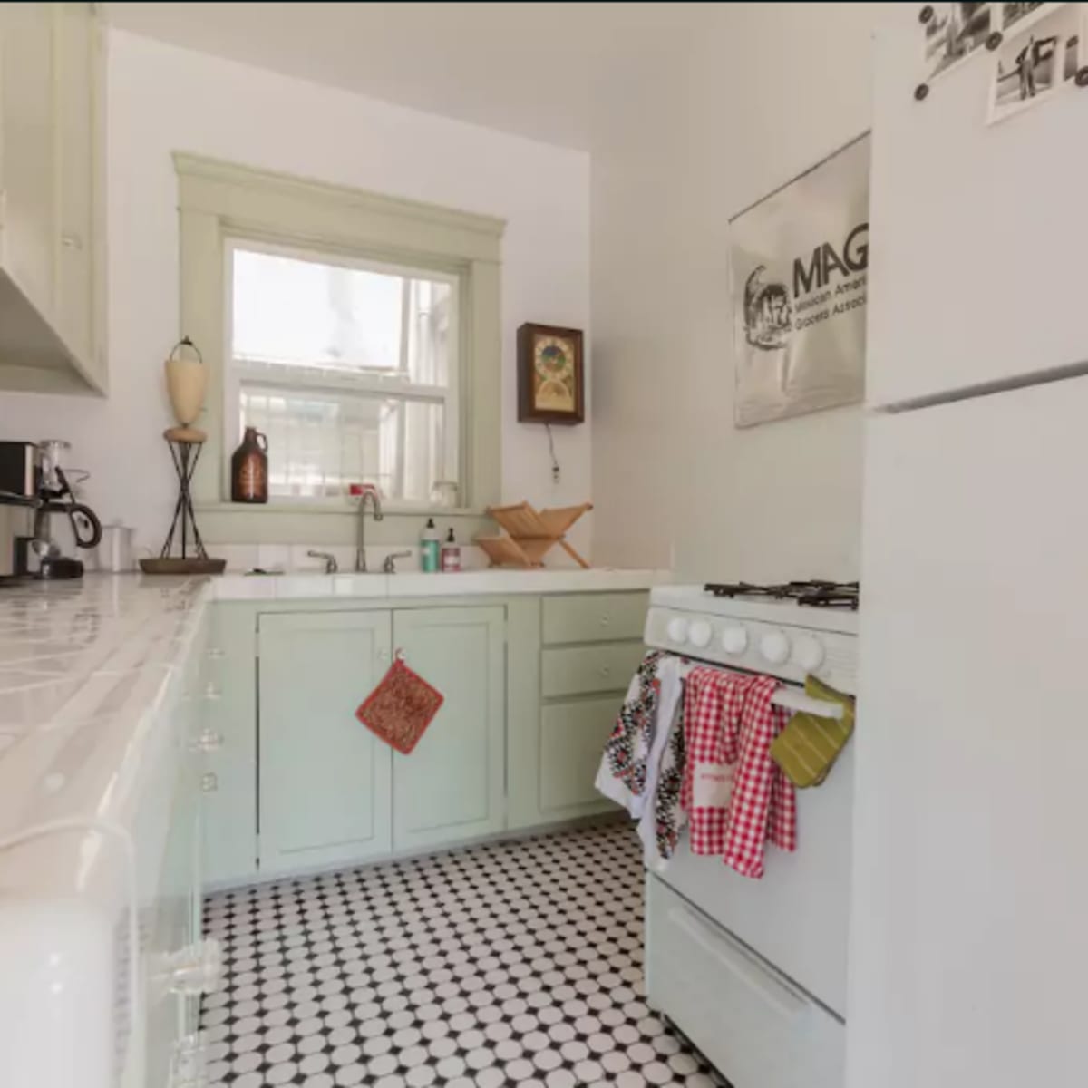 A small kitchen featuring light green cabinets, a white gas stove, framed wall art, and black-and-white tiled flooring.