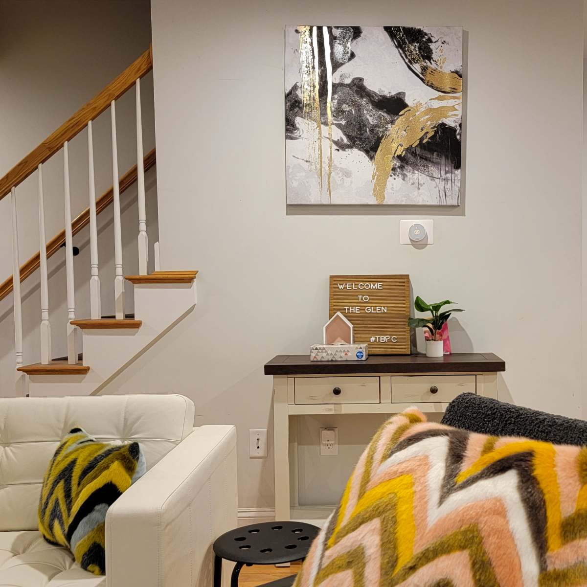 A cozy living room featuring a white couch, a small table with a welcome sign, a colorful abstract painting on the wall, and stairs leading to an upper level.