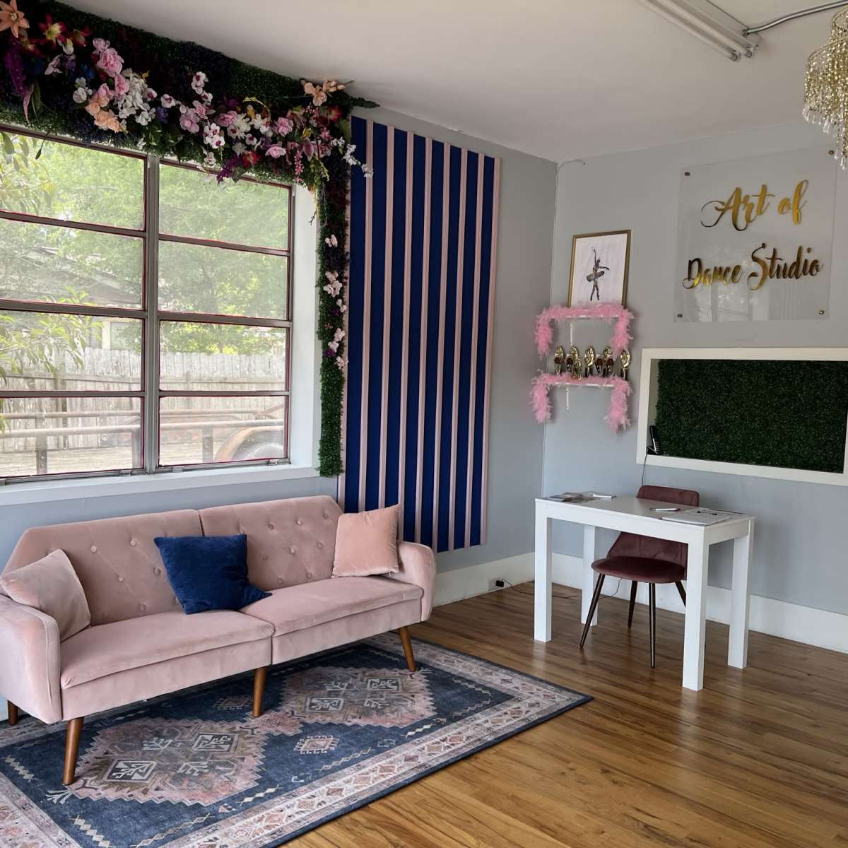 A cozy studio space featuring a pink sofa, a small table with a chair, and decorative elements on the walls, including a floral arrangement and a mirror.