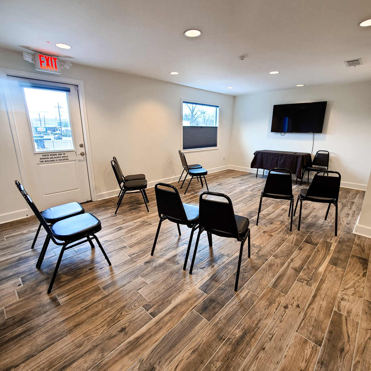 A small, bright room features a row of six black chairs arranged in a circle around a table with a television mounted on the wall.