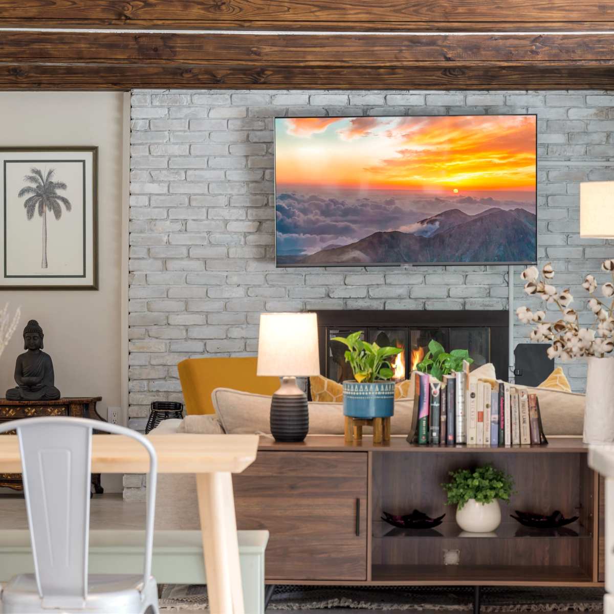 A cozy living room features a mounted television displaying a sunset over mountains, with a yellow couch, a coffee table, and a decorative bookshelf.